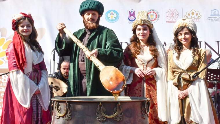 Yeşil kostümlü bir adam, geleneksel kıyafetli üç kadının yanında büyük bir kazanın başında duruyor.
