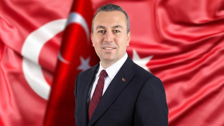Takım elbiseli bir kişi, Türk bayrağı önünde kameraya bakarak duruyor.