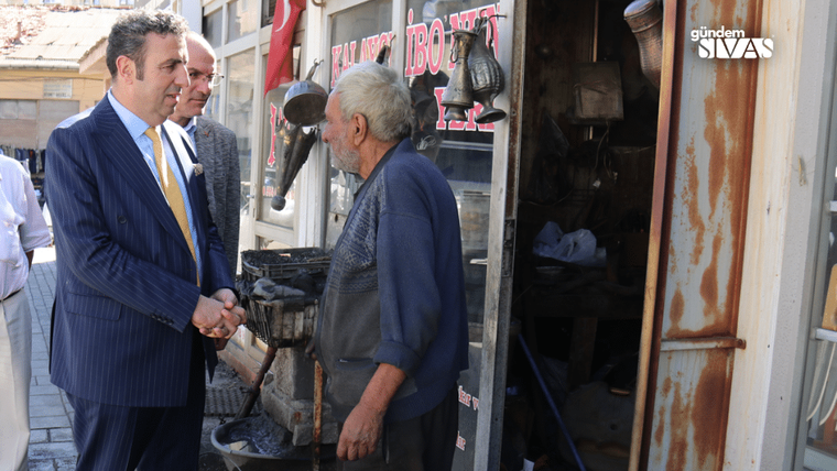 Takım elbiseli bir kişi, dükkân önünde yaşlı bir erkekle el sıkışıyor; kapıda ayakkabı malzemeleri görülüyor.