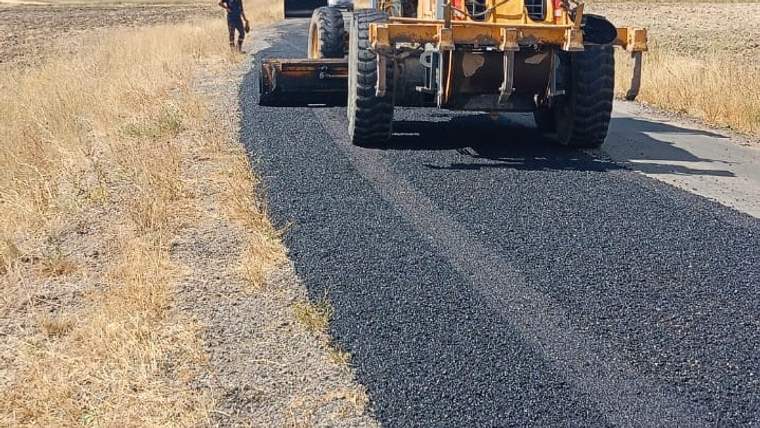 Kırsal bir yolda asfalt serim makinesi ilerlerken, yolun bir bölümüne siyah asfalt dökülmüş görünüyor.