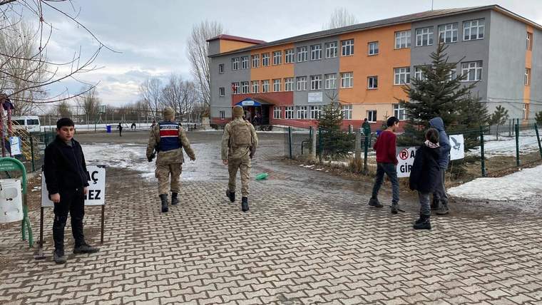 Okul binası önündeki taş döşeli alanda iki üniformalı kişi ve birkaç genç görülüyor.