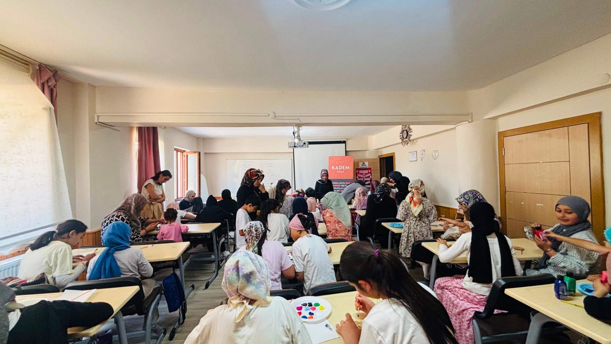 Sınıf ortamında, çeşitli yaşlarda kız çocukları ve kadınlar etkinlik yapıyor.