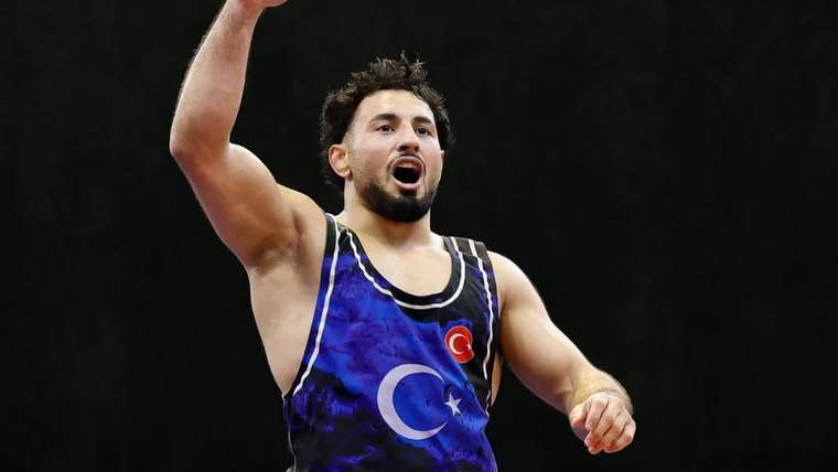 Mavi güreş formasındaki sporcu, minderde bir kolunu havaya kaldırarak ilerlerken arka planda skor ekranı görülüyor.
