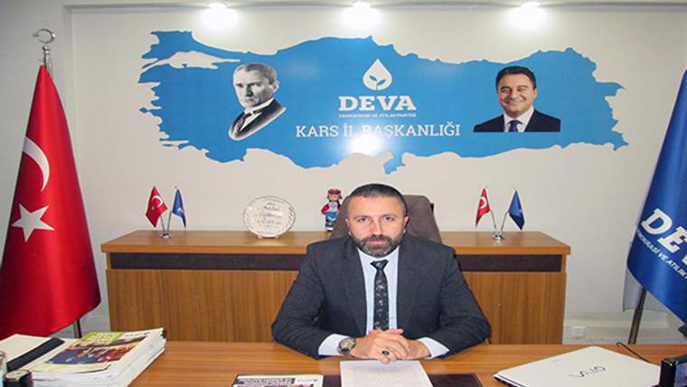 Takım elbiseli bir kişi, arkasında “DEVA Kars İl Başkanlığı” yazılı pano bulunan masada oturuyor.