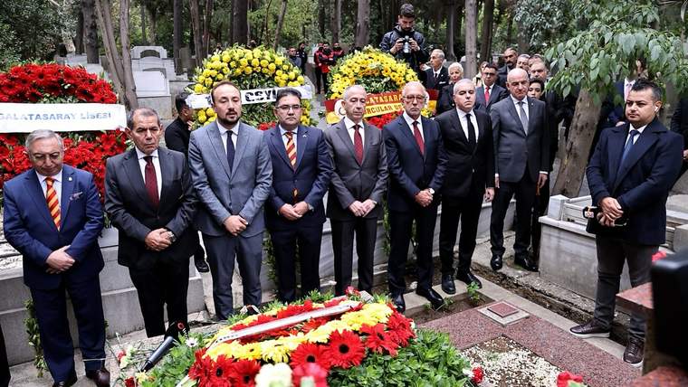 Mezar taşları ve çiçeklerle çevrili alanda, takım elbiseli bir grup erkek çelenklerin önünde duruyor.
