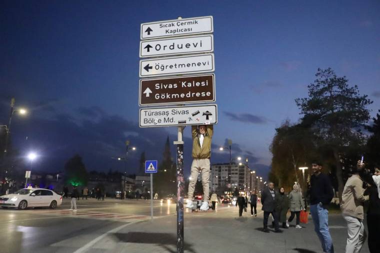 Bir genç, trafik tabelasına asılarak duruyor; arka planda yürüyen insanlar ve sokak aydınlatmaları görünmekte.