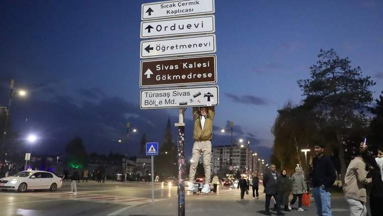 Bir genç, trafik tabelasına asılarak duruyor; arka planda yürüyen insanlar ve sokak aydınlatmaları görünmekte.