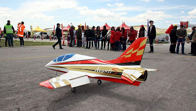 Küçük bir model uçak, pistte duruyor; arka planda kalabalık ve etkinlik alanı görünmekte.