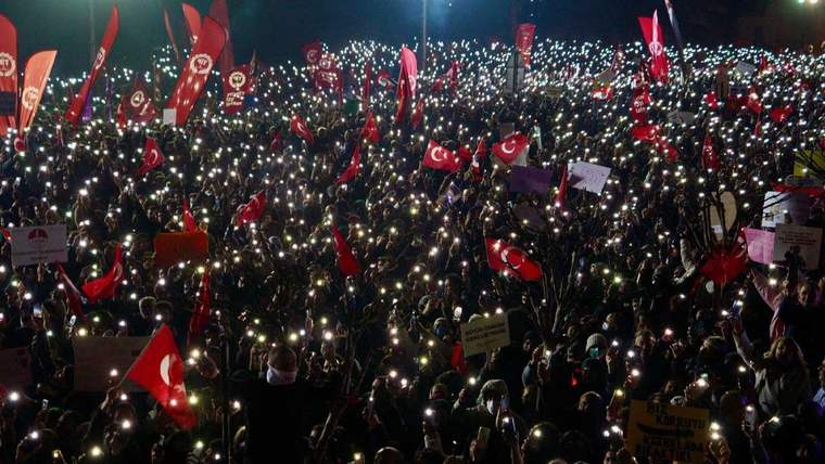 Karanlık bir alanda, ellerinde ışık tutan telefonlar ve Türk bayraklarıyla dolu kalabalık bir miting.