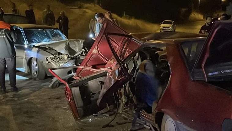 Gece yolunda ağır hasar görmüş kırmızı otomobilin yanında, önü ezilmiş başka bir otomobil duruyor.