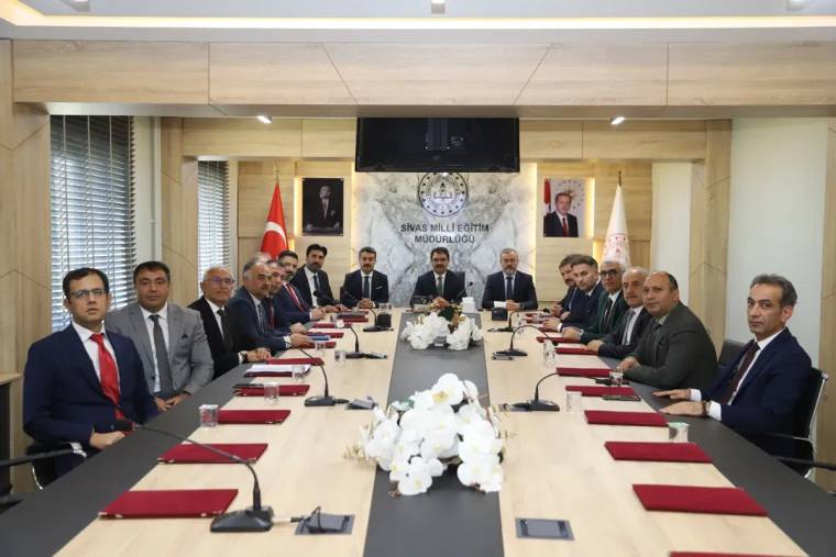 Toplantı salonunda uzun masa çevresinde oturan kişiler, masadaki mikrofon ve çiçeklerle birlikte görülüyor.