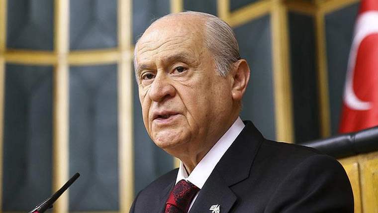 Mikrofon önünde konuşan, takım elbiseli ve kravatlı bir kişi; arka planda Türkiye bayrağı görülüyor.