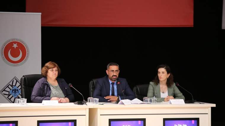 Panel masasında oturan üç kişi, önlerindeki mikrofon ve belgelerle birlikte izleyicilere dönük duruyor.