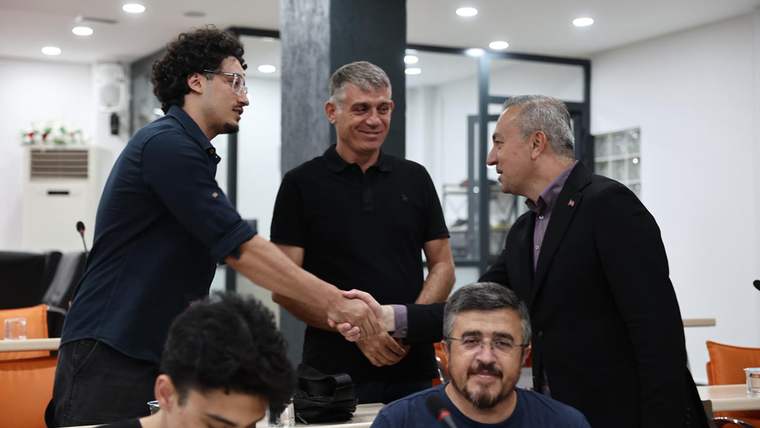 Bir grup erkek, bir toplantı salonunda el sıkışıyor; arka planda başka kişiler ve masalar var.