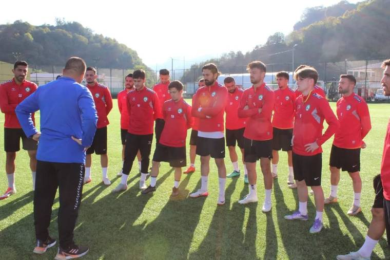 Kırmızı formalar giyen futbolcular, antrenman sahasında mavi üstlü bir kişinin karşısında toplu halde duruyor.