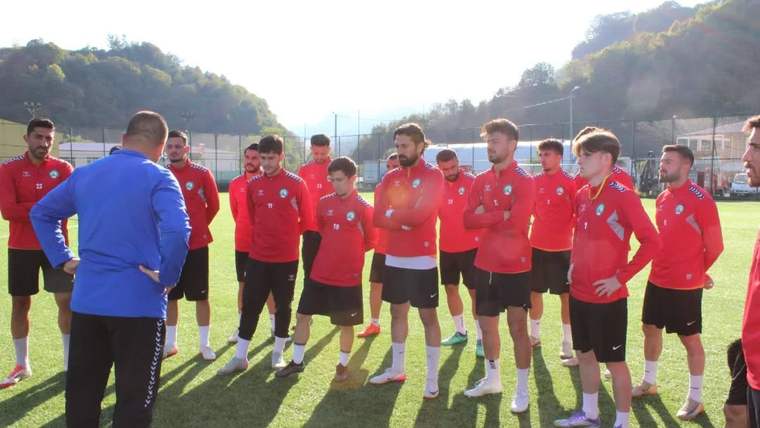 Kırmızı formalar giyen futbolcular, antrenman sahasında mavi üstlü bir kişinin karşısında toplu halde duruyor.