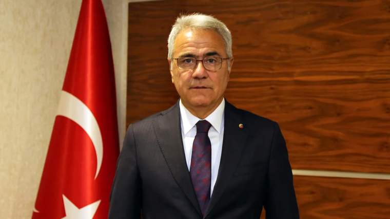 Kırmızı-beyaz Türk bayrağının önünde, takım elbise giymiş bir kişi poz veriyor.