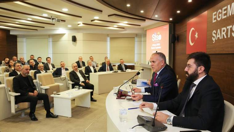 Toplantı salonunda dinleyicilere dönük oturan kişiler ve masada konuşan iki kişi görülüyor.