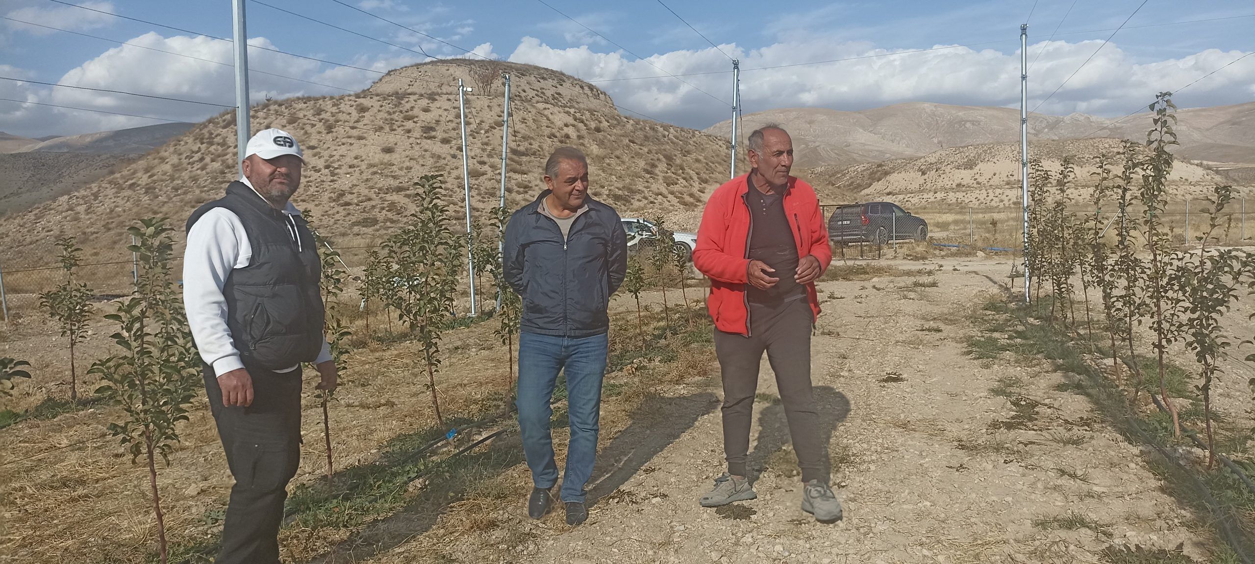 Üç kişi, elma bahçesinin yanında duruyor; arka planda dağlar ve ağaçlar var.