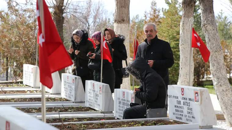 Türk bayraklarıyla çevrili mezarların önünde oturan ve dua eden insanlar.