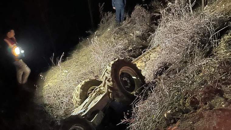 Gece karanlığında, eğimli kayalık arazide ters dönmüş bir traktör ve çevresinde duran iki kişi görülüyor.