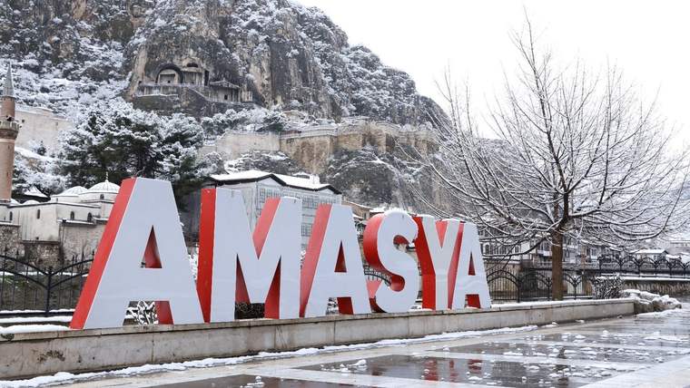 Kar yağışı sonrası Amasya yazılı şehir tabelası, kayalık yamaç ve karla kaplı ağaçlar önünde görülüyor.