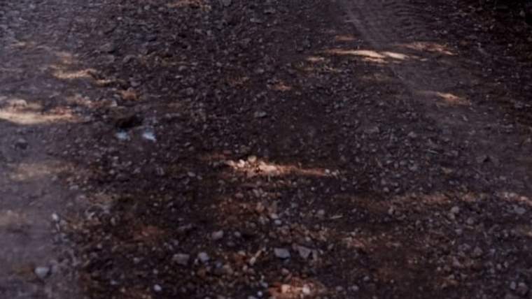 Yakın planda, koyu renkli mıcır ve taşlarla kaplı asfalt yol yüzeyi görülüyor.
