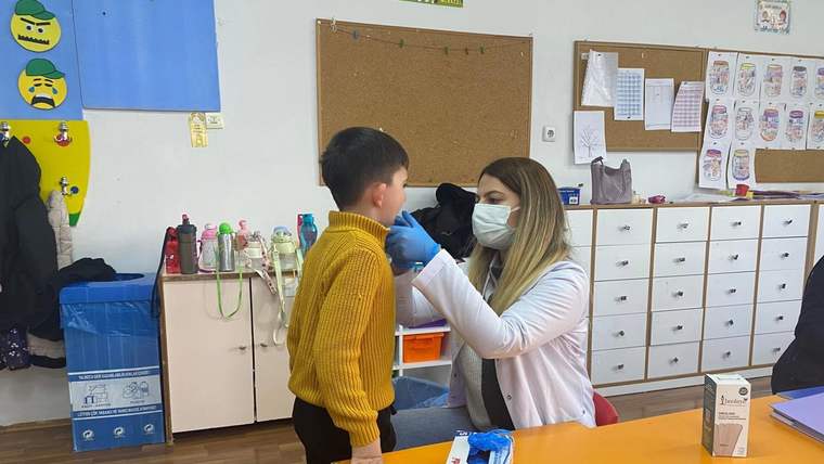 Beyaz önlüklü ve maskeli bir kişi, sınıfta ayakta duran çocuğun ağzını mavi eldivenle kontrol ediyor.