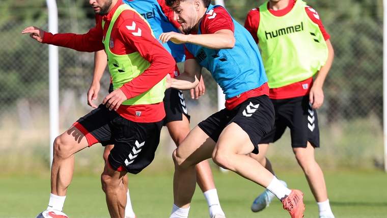 Çim sahada forma ve antrenman yeleği giyen futbolcular, top için ikili mücadele ediyor.