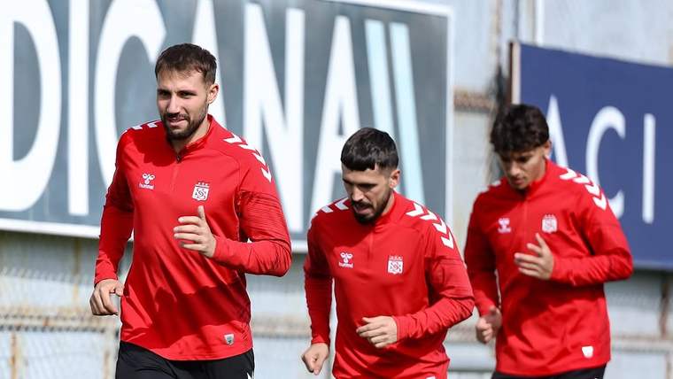 Kırmızı formalar ve siyah şortlar giyen üç futbolcu, antrenman sahasında yan yana koşuyor.