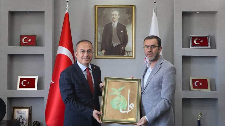 Türk bayrağı ve Atatürk portresiyle süslü bir ofiste, iki kişi bir tabloyu tutuyor.