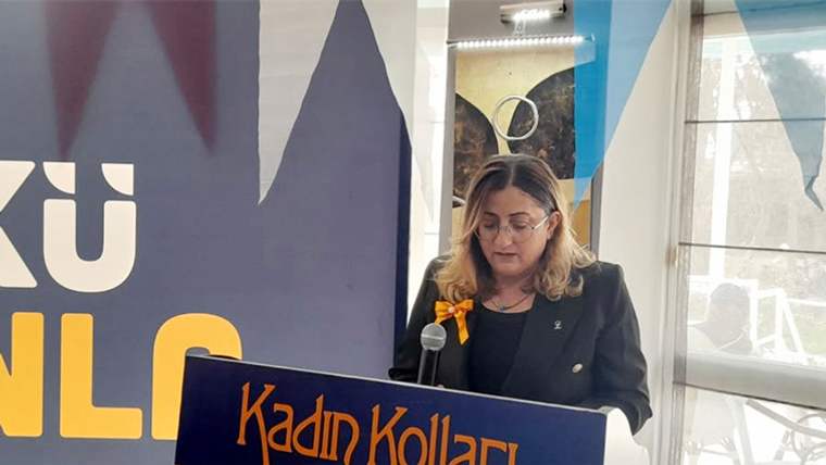 Kürsüde mikrofonla konuşan gözlüklü bir kadın, üzerinde “Kadın Kolları” yazan afişin önünde duruyor.