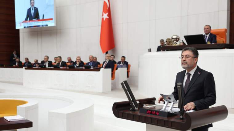 Takım elbiseli bir kişi, meclis salonunda kürsüde konuşurken arka planda Türk bayrağı ve ekran görülüyor.