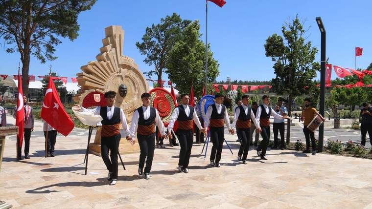 Türk bayraklarıyla süslenmiş alanda, geleneksel kıyafetli erkekler taş anıtın önünde yürürken görülüyor.