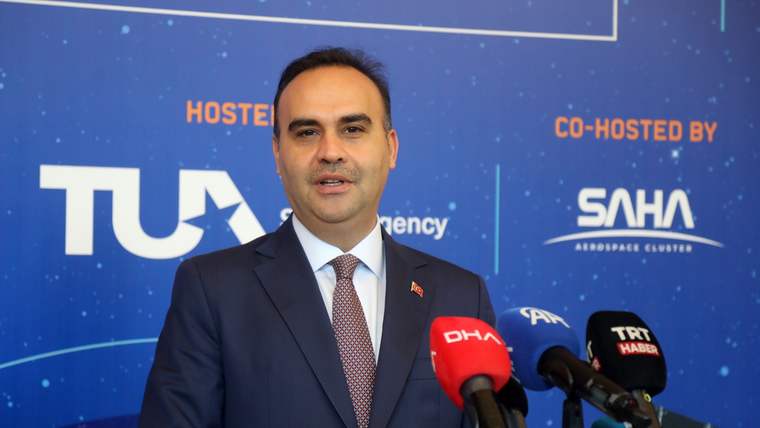 Takım elbiseli bir kişi, üzerinde TUA ve SAHA logoları bulunan panonun önünde mikrofonlara konuşuyor.
