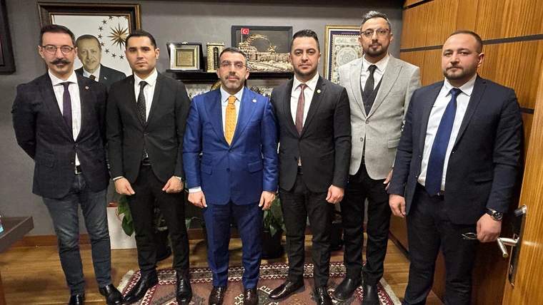Bir odada takım elbiseli altı erkek, duvarındaki çerçeveler ve halı önünde yan yana duruyor.