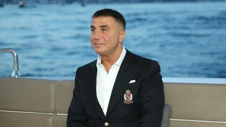 Siyah ceket giymiş bir adam, gülümseyerek oturuyor. Arka planda su görünmekte.