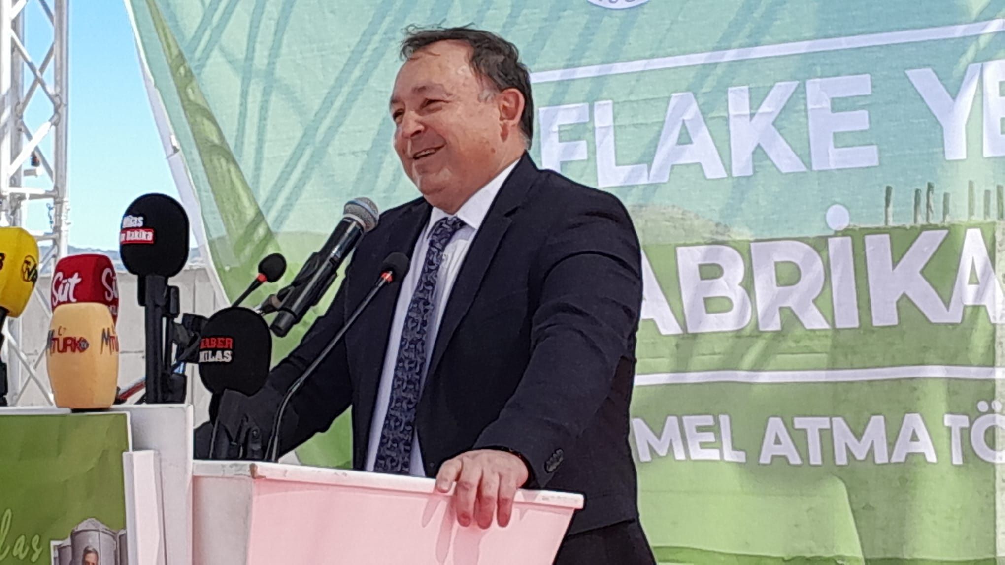 Kürsüde konuşan takım elbiseli bir kişi, önündeki mikrofonlar ve arkasındaki Flake Yem Fabrikası pankartıyla görülüyor.