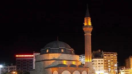 Gece aydınlatılan cami, kubbesi ve tek minaresiyle şehir merkezindeki binaların arasında görülüyor.