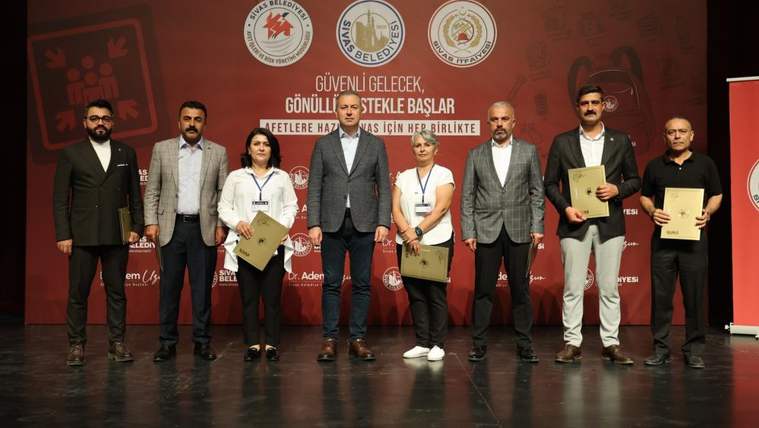 Kırmızı afiş önünde duran yedi kişi, ellerindeki belgelerle sahnede yan yana poz veriyor.
