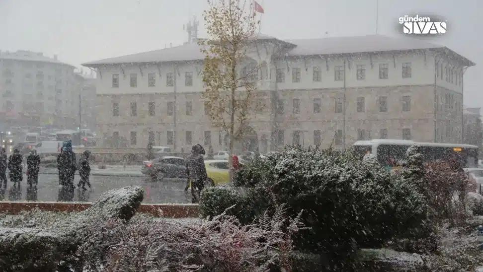 Kar yağışı altında yürüyen insanlar ve karla kaplı bitkiler, arka planda bir bina görünmekte.