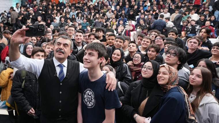 Bir grup öğrenci, bir kişiyle birlikte selfie çekiyor. Kalabalık bir arka planda gençler gülümseyerek poz veriyor.