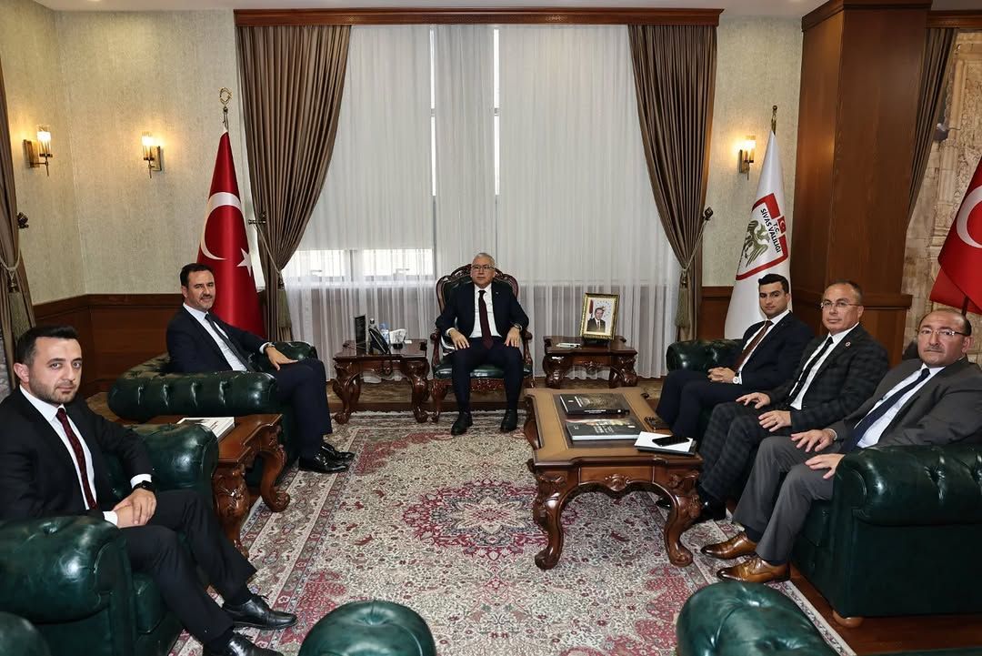 Türk bayrakları ve kurum flamaları bulunan salonda, takım elbiseli altı kişi koltuklarda oturuyor.