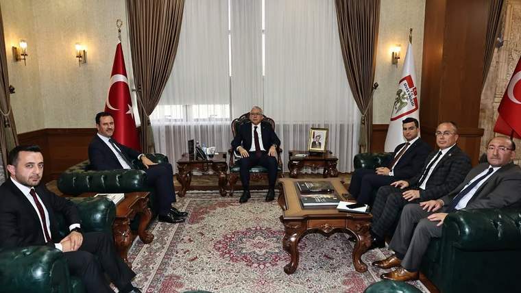 Türk bayrakları ve kurum flamaları bulunan salonda, takım elbiseli altı kişi koltuklarda oturuyor.