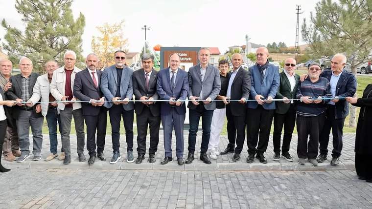 Ağaçların bulunduğu alanda bir grup kişi, ellerindeki makaslarla kurdele kesme törenine katılıyor.