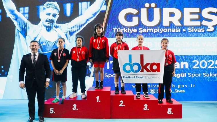 Beş çocuk, madalyalarıyla güreş şampiyonası kürsüsünde duruyor; yanlarında bir yetişkin ve sponsor panosu bulunuyor.