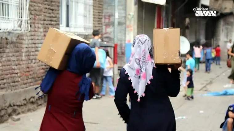 Sokakta başlarında karton kutular taşıyan iki kadın, arka planda çocuklar ve binalar görülüyor.
