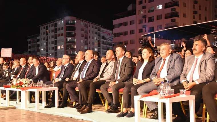 Gece düzenlenen açık hava etkinliğinde, takım elbiseli kişiler yan yana oturuyor; önlerinde çiçekli masalar bulunuyor.