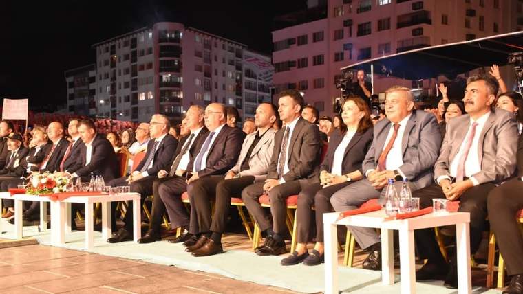 Gece düzenlenen açık hava etkinliğinde, takım elbiseli kişiler yan yana oturuyor; önlerinde çiçekli masalar bulunuyor.