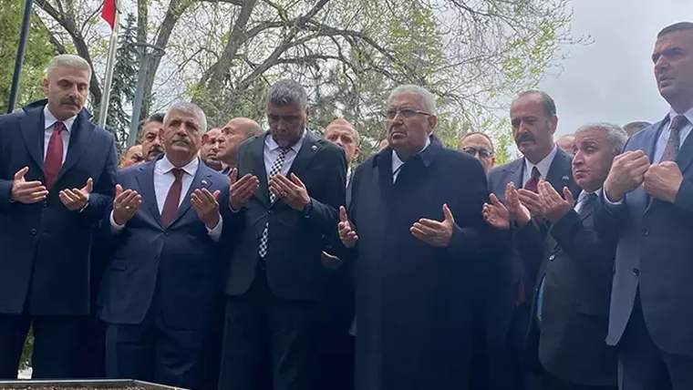 Ağaçlar arasındaki mezarlıkta, takım elbiseli bir grup erkek ellerini dua eder gibi açarak duruyor.